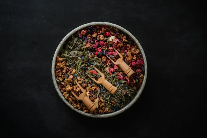 Bowl of loose herbal tea blends with three small wooden scoops on a dark background