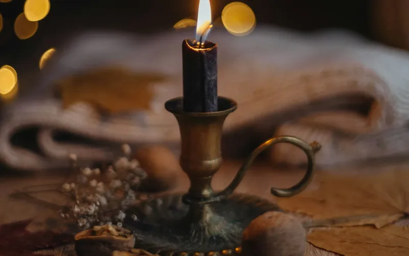 A lit candle in a brass holder on a table with autumn leaves and a soft knitted throw behind