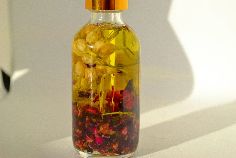 Glass bottle of golden oil surrounded by jasmine buds and red rose petals