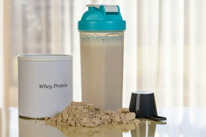 A tub of whey protein beside a filled shaker bottle, a scoop and a pile of powder