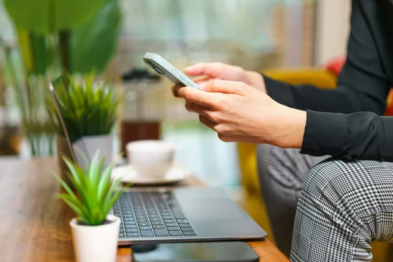Hands holding a smartphone beside a laptop and a cup of coffee