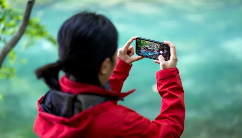 Traveller in a red jacket photographing a turquoise river with a smartphone