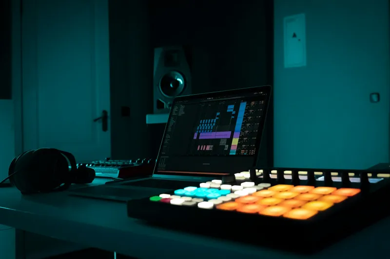 A laptop running music software beside a lit drum pad controller and headphones in a dim studio