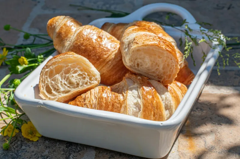 Golden butter croissants, one cut open, in a white ceramic dish on a sunny stone surface