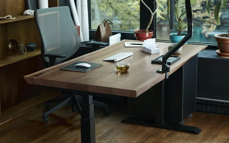 A wooden standing desk with a monitor arm, keyboard and a cup of tea in a home office by a window