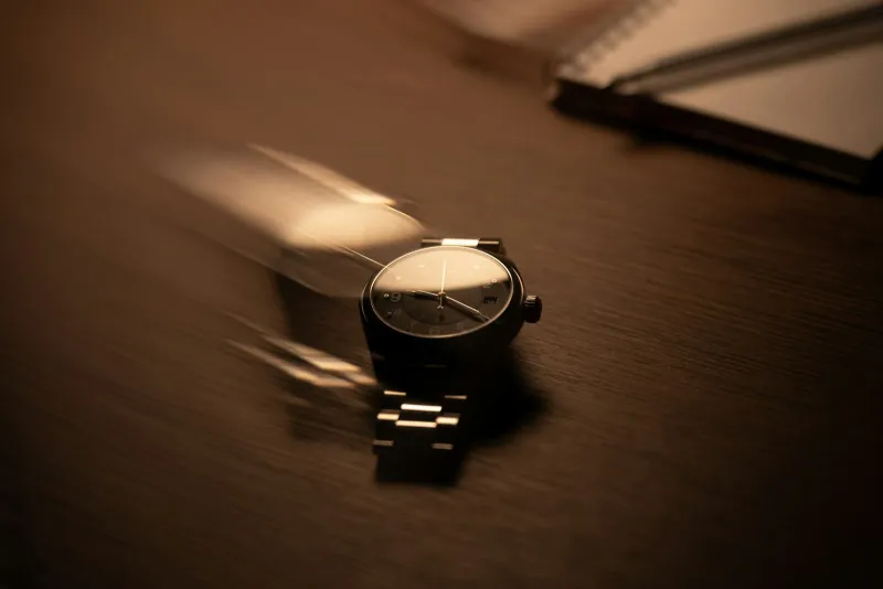 Black wristwatch with a metal bracelet on a dark wooden desk beside a notebook
