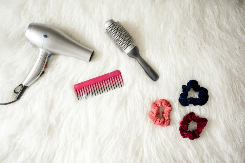 Silver hair dryer, round brush, pink comb and velvet scrunchies on a white rug