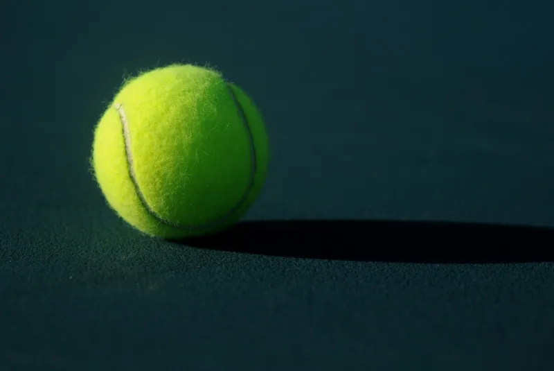 A yellow tennis ball casting a long shadow on a green hard court