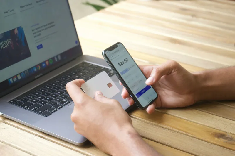 Person paying online with a card and phone beside a laptop